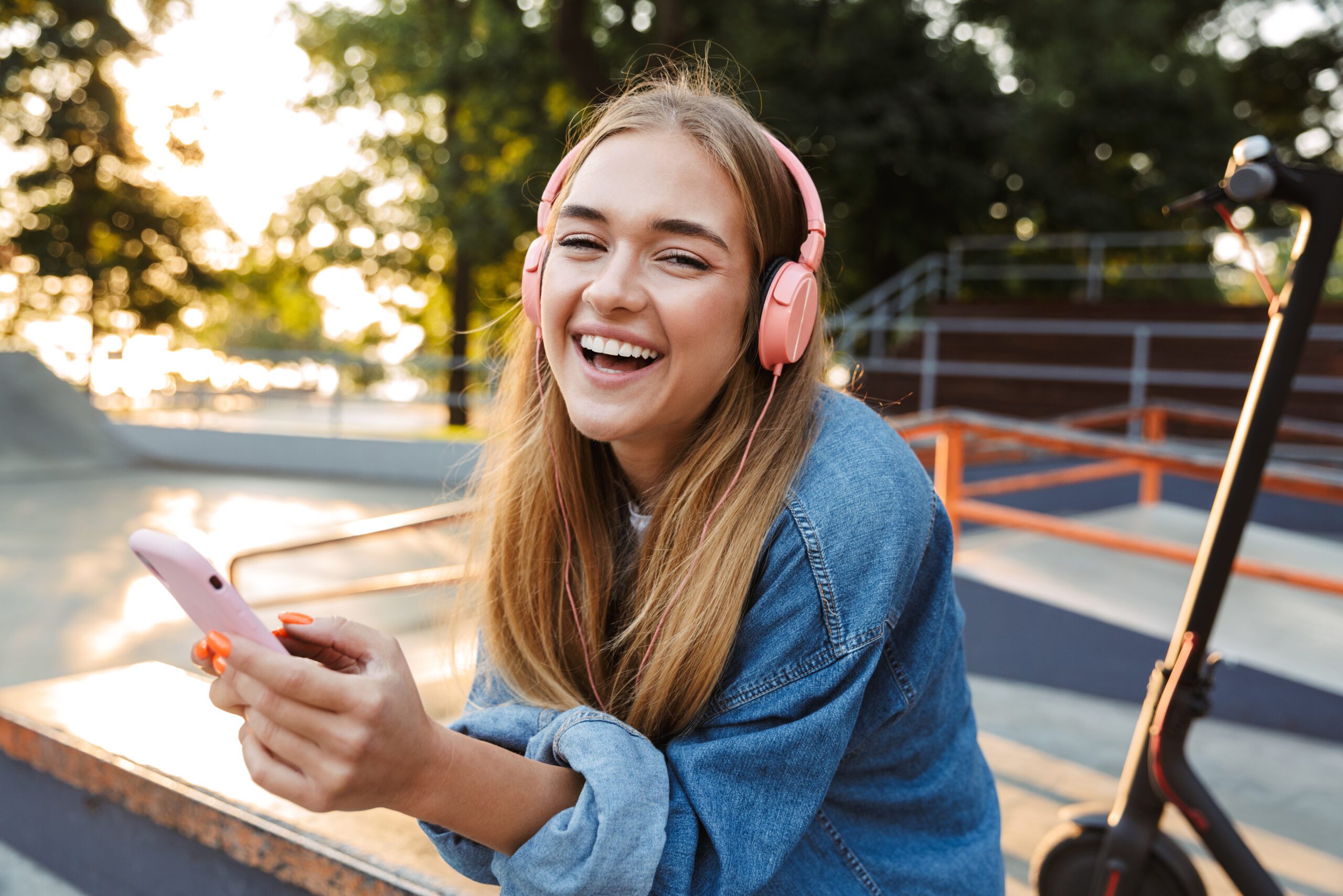 Image of a laughing cheery smiling young teenage girl outside in park listening music with headphones holding mobile phone showing tongue.
