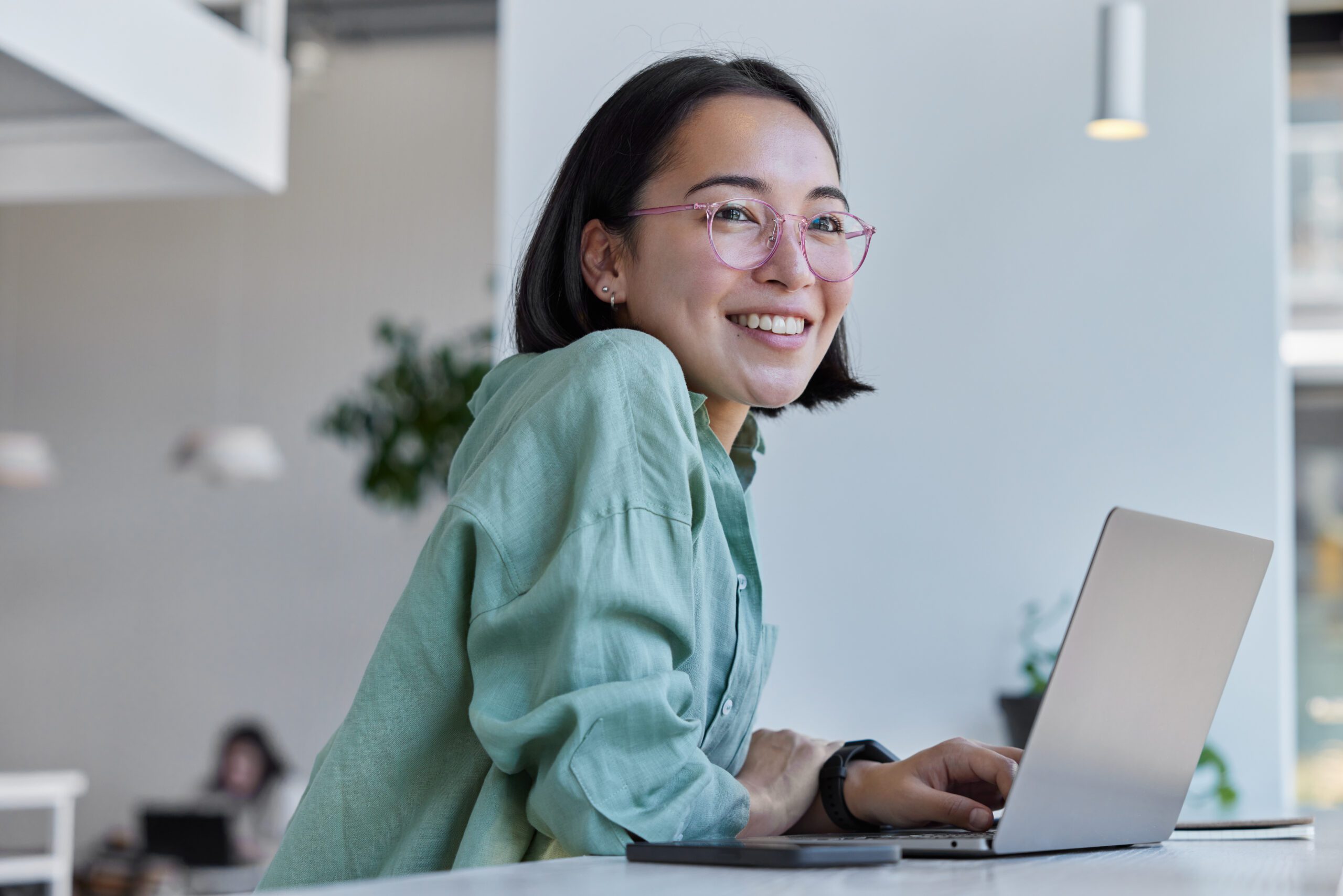 Pretty cheerful Asian woman in eyeglasses and casual clothes browses laptop computer connected to 4g internet updates software uses modern technologies poses in cafeteria looks gladfully away