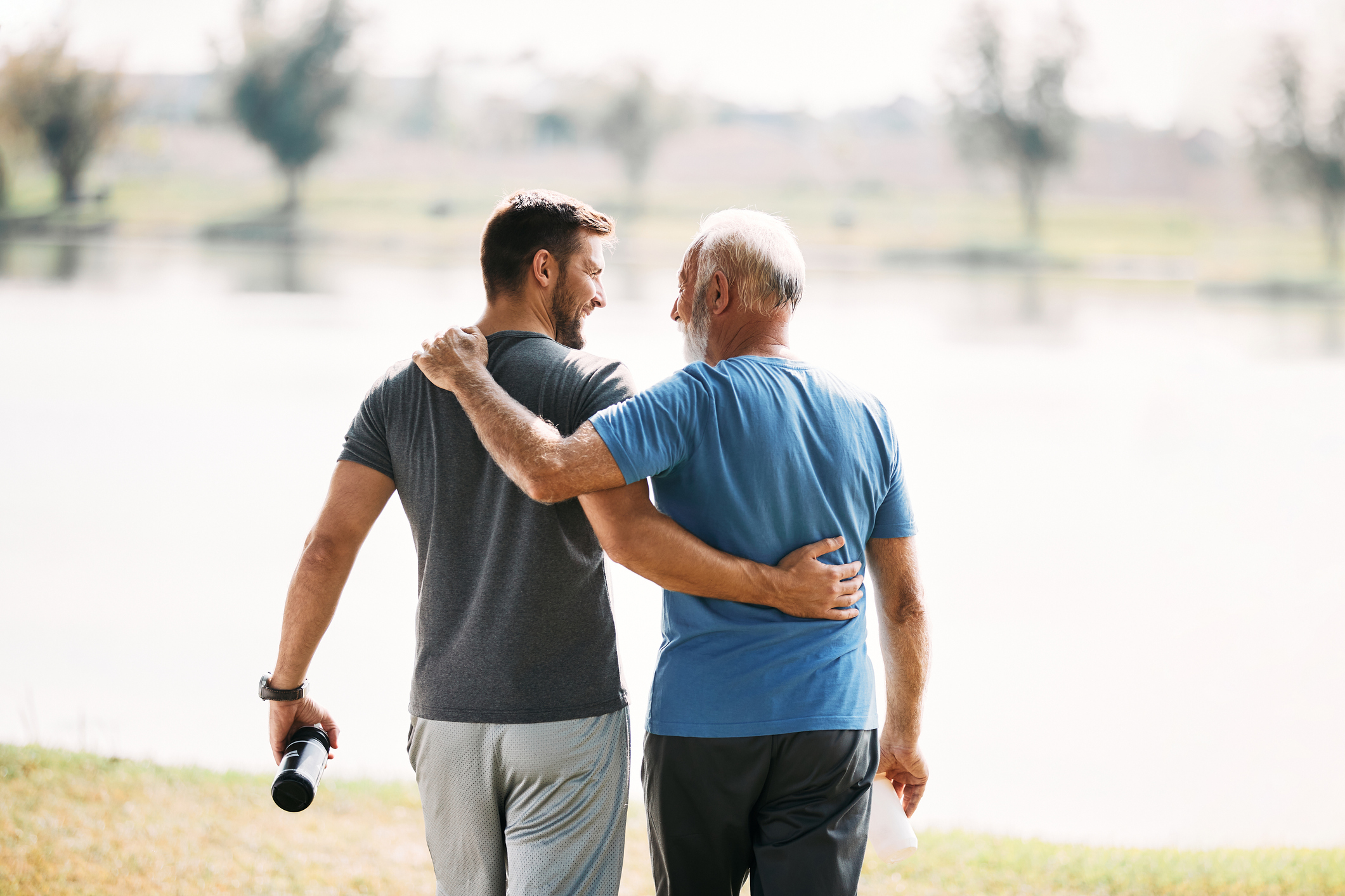 Back view of happy athletic man and his senior father walking embraced while exercising in nature.